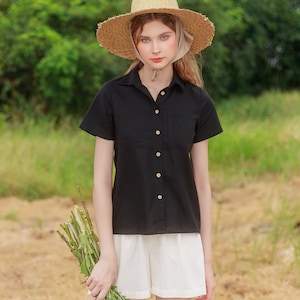 May include: A woman wearing a black collared shirt with short sleeves and white shorts. She is wearing a straw hat with a light brown ribbon. She is holding a bouquet of green flowers.