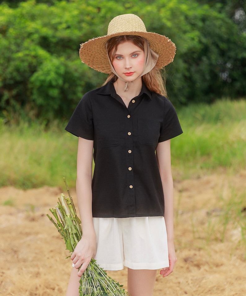 May include: A woman wearing a black collared shirt with short sleeves and white shorts. She is wearing a straw hat with a light brown ribbon. She is holding a bouquet of green flowers.