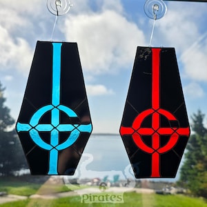 May include: Two stained glass suncatchers in a window, each in a coffin shape. One features a blue design, the other red, both against a black background. The design is a cross within a circle, with a vertical line.