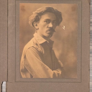 Könnte beinhalten: Sepiafarbenes Porträt eines Mannes mit Schnurrbart, der ein Hemd mit Kragen trägt und mit verschränkten Armen posiert. Das Foto ist in einem braunen Papierrahmen montiert, was eine Vintage-Ästhetik suggeriert. Das Bild ist leicht verblasst und verleiht ihm einen antiken Charme.