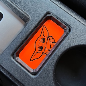 May include: A black car interior with a phone holder featuring a cartoon image of a character from the Star Wars franchise. The phone holder is made of clear plastic with an orange background.