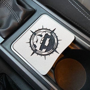 May include: A silver metal cup holder with a black and white compass design. The compass design features a stylized truck with a sun and moon in the background.