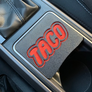 May include: A black car cup holder with a red "TACO" graphic. The graphic is in a bold, stylized font.