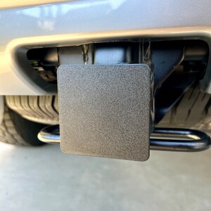 May include: A black, textured trailer hitch cover is installed on a vehicle. The cover is rectangular and fits snugly into the hitch receiver. The hitch features a pair of curved safety chain loops.