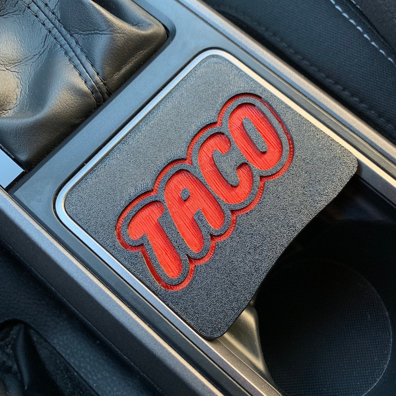 May include: A black car cup holder with a red "TACO" graphic. The graphic is in a bold, stylized font.
