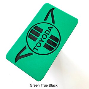 K&ouml;nnte beinhalten: Ein rechteckiger, gr&uuml;ner Gegenstand mit dem schwarzen Wort "TOYODA". Der Text befindet sich in einem schwarzen Kreis mit schwarzen Linien. Die Oberfl&auml;che ist strukturiert und der Text "Green True Black" steht unten.