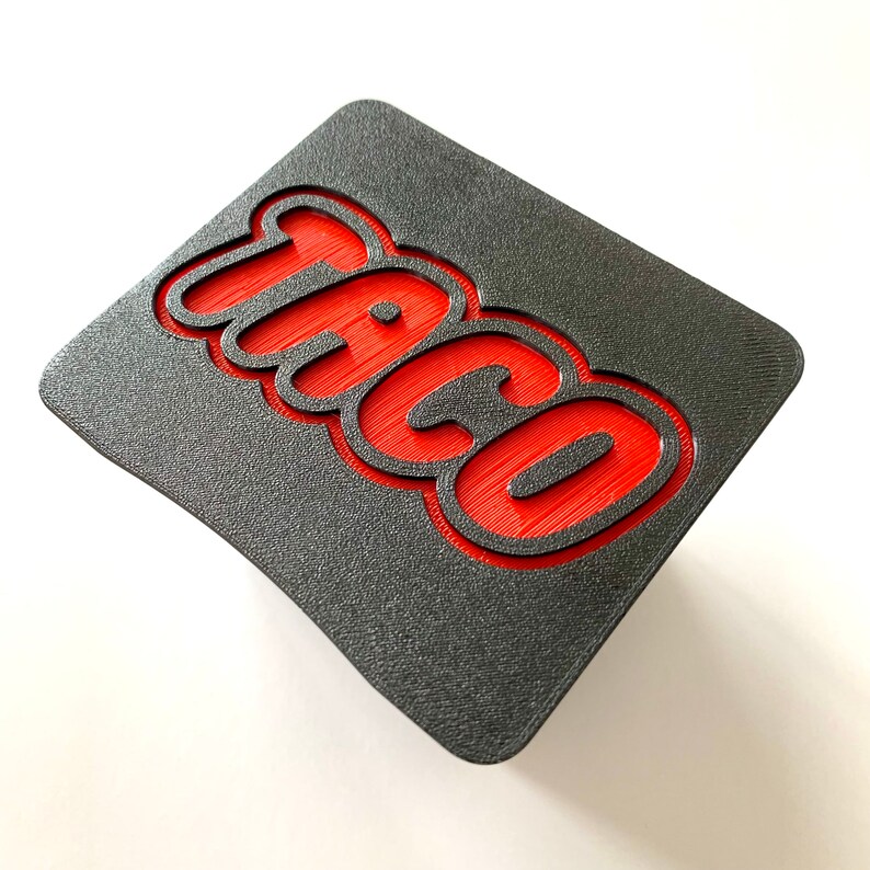 May include: A black and red 3D printed sign with the word "Taco" in red raised letters.