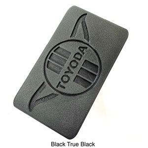 K&ouml;nnte beinhalten: Ein rechteckiger, schwarzer Gegenstand mit dem Wort "TOYODA" in einem kreisf&ouml;rmigen Emblem. Der Gegenstand hat eine strukturierte Oberfl&auml;che und ein Design auf jeder Seite des Emblems. Der Text "Black True Black" steht unten.