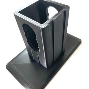 K&ouml;nnte beinhalten: Ein schwarzer, 3D-gedruckter Halter mit rechteckiger Basis und einem gekerbten Fach mit offenem Deckel. Der Text "Universal Notched Design" ist unten aufgedruckt. Der Halter ist zur Aufnahme eines Gegenstandes bestimmt.