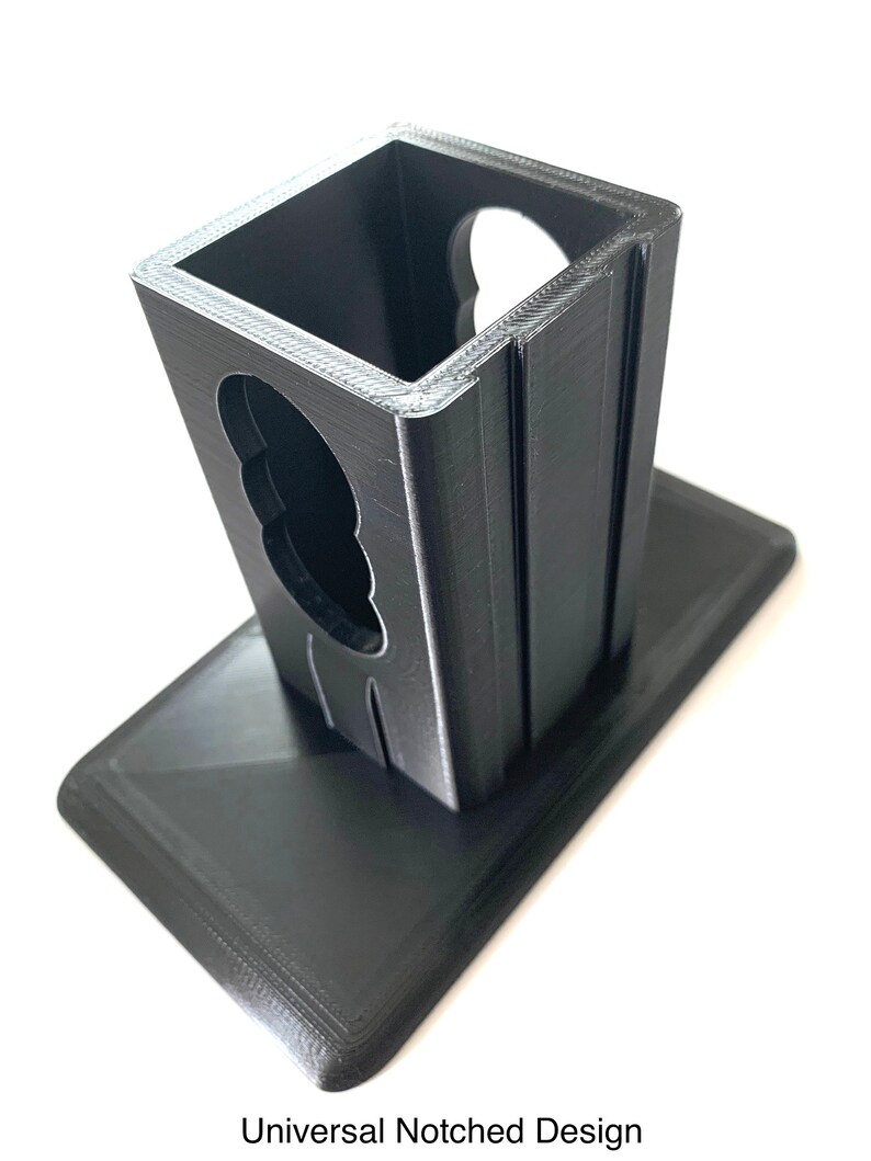 K&ouml;nnte beinhalten: Ein schwarzer, 3D-gedruckter Halter mit rechteckiger Basis und einem gekerbten Fach mit offenem Deckel. Der Text "Universal Notched Design" ist unten aufgedruckt. Der Halter ist zur Aufnahme eines Gegenstandes bestimmt.