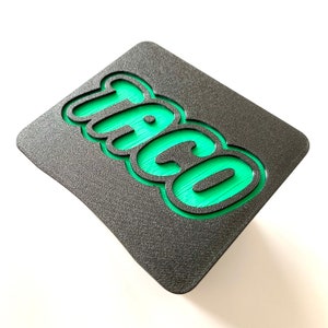 May include: A black and green sign with the word "Taco" in a 3D, bubble-like font.