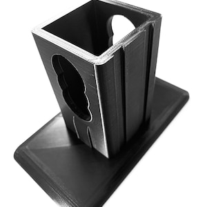 May include: A black 3D printed phone stand with a notched design. The stand has a rectangular base and a square top with a cutout for a phone.