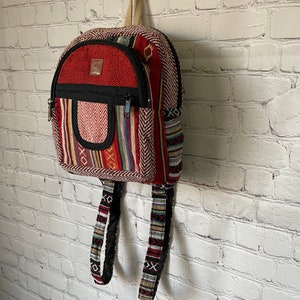 K&ouml;nnte beinhalten: Ein kleiner, farbenfroher Rucksack mit roter Oberseite und einer schwarzen Rei&szlig;verschlusstasche. Der Rucksack weist eine Vielzahl von Webmustern in Rot, Schwarz und anderen Farben auf. Die Tr&auml;ger und der Griff sind beige.