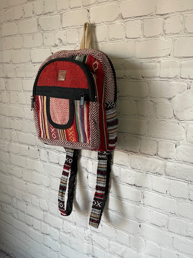 K&ouml;nnte beinhalten: Ein kleiner, farbenfroher Rucksack mit roter Oberseite und einer schwarzen Rei&szlig;verschlusstasche. Der Rucksack weist eine Vielzahl von Webmustern in Rot, Schwarz und anderen Farben auf. Die Tr&auml;ger und der Griff sind beige.