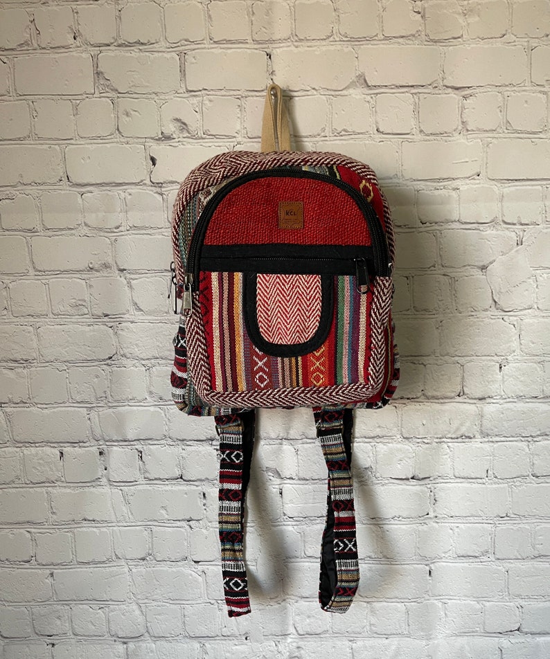 May include: A small, colorful backpack with a red top and a patterned front. The backpack features a zippered main compartment, a front pocket, and adjustable straps. The design incorporates various colors and geometric patterns, with a neutral-colored handle.