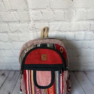 K&ouml;nnte beinhalten: Ein kleiner, farbenfroher Rucksack mit roter Oberseite und einer Vordertasche mit Zickzackmuster. Die Tasche hat einen schwarzen Rei&szlig;verschluss und einen kleinen Griff. Der Rucksack hat ein gestreiftes Muster in verschiedenen Farben.