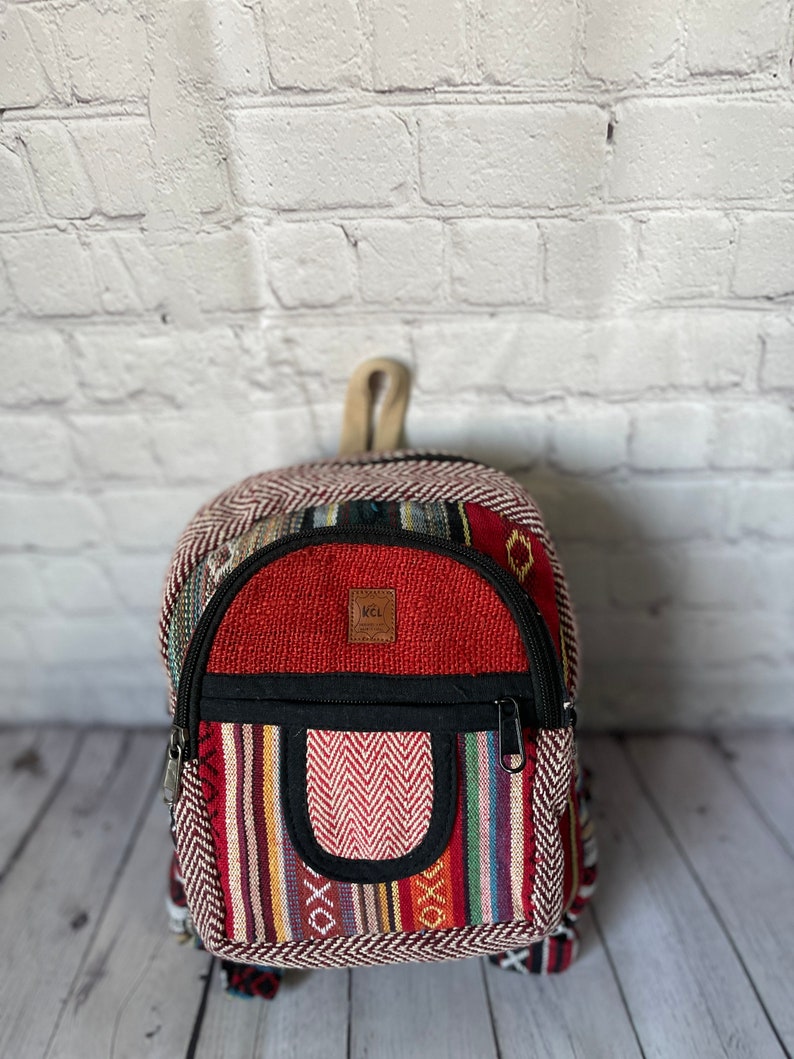 K&ouml;nnte beinhalten: Ein kleiner, farbenfroher Rucksack mit roter Oberseite und einer Vordertasche mit Zickzackmuster. Die Tasche hat einen schwarzen Rei&szlig;verschluss und einen kleinen Griff. Der Rucksack hat ein gestreiftes Muster in verschiedenen Farben.