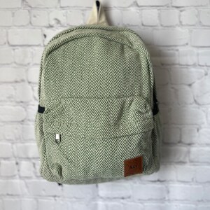 May include: Green and white herringbone pattern backpack with a small front pocket and a zipper closure. The backpack has a cream-colored handle and a brown leather tag with text. The bag is hanging on a white brick wall.
