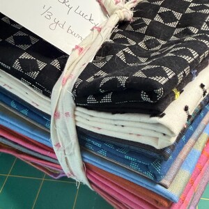 Op de afbeelding: Een stapel gevouwen stoffen vastgebonden met een witte stoffen strook. De bovenste stof is zwart met een wit geometrisch patroon. Andere lagen bevatten crèmekleurige, blauwe, roze en oranje textiel. Een wit label vermeldt Warp + Weft Ohh Lucky Lucky 1/3 yd bundle.