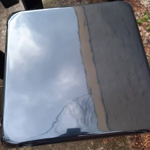 May include: A black, glossy, square-shaped stool with rounded edges. The stool is made of a smooth, hard material and has a reflective surface.