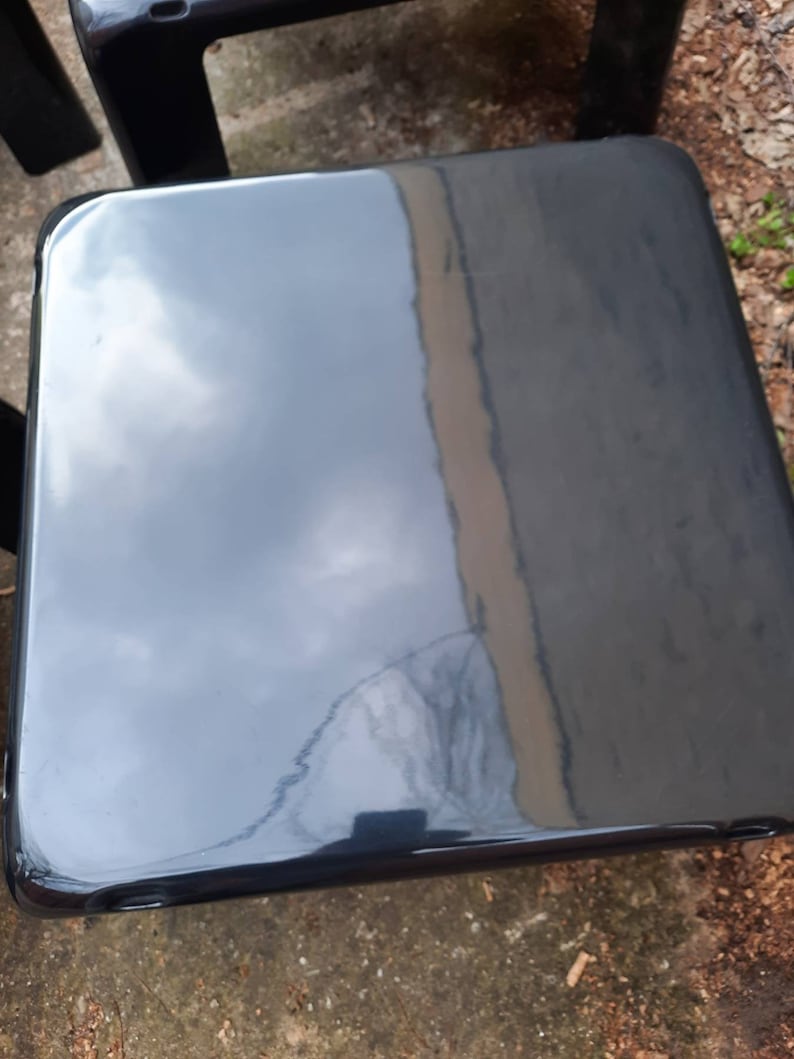 May include: A black, glossy, square-shaped stool with rounded edges. The stool is made of a smooth, hard material and has a reflective surface.