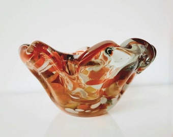 May include: A clear glass ashtray with a wavy, organic shape. The ashtray has a brown, white, and orange swirl pattern.