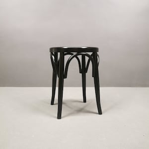 May include: A black wooden stool with a round seat and curved back supports. The stool has four legs and a dark finish. The design is simple and classic, suitable for various interior styles.