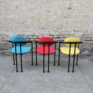 May include: Three chairs with black metal frames and different colored fabric seats. The chairs are lined up in a row against a brick wall. The chair on the left has a blue seat, the middle chair has a red seat, and the chair on the right has a yellow seat.