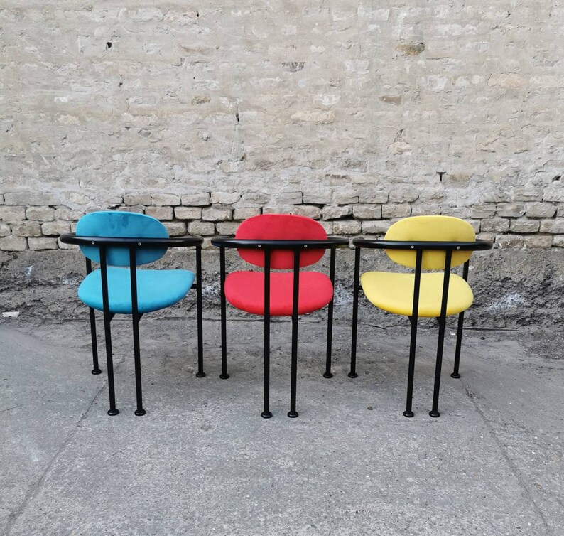 May include: Three chairs with black metal frames and different colored fabric seats. The chairs are lined up in a row against a brick wall. The chair on the left has a blue seat, the middle chair has a red seat, and the chair on the right has a yellow seat.