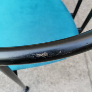 May include: Close-up of a black metal chair with a teal blue fabric seat. The chair has a circular back and a single leg in the foreground. The black paint on the chair is chipped and worn.