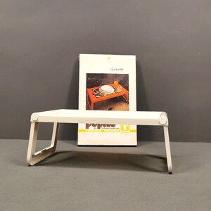 Puede incluir: Mesa plegable blanca con una superficie blanca y patas de metal blanco. La mesa está plegada y se encuentra sobre una superficie gris. Una caja con el texto "Pop Up" está detrás de la mesa.