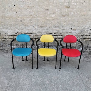 May include: Three chairs with black metal frames and colorful fabric seats. The chairs are arranged in a row against a brick wall. The chair on the left has a blue seat, the middle chair has a yellow seat, and the chair on the right has a red seat.