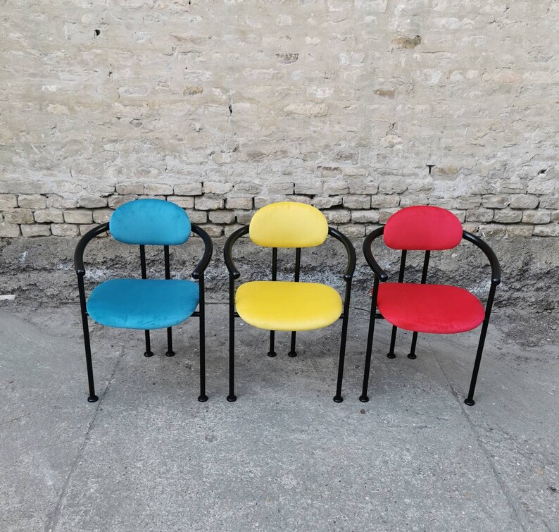 May include: Three chairs with black metal frames and colorful fabric seats. The chairs are arranged in a row against a brick wall. The chair on the left has a blue seat, the middle chair has a yellow seat, and the chair on the right has a red seat.
