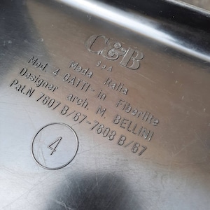 May include: Close-up of a black plastic label with white text. The label reads "C&B SpA Mod. 4 GATTI-in Fiberlite Meda Italia Designer arch. M. BELLINI Pat.N 7607 B/67-7608 B/67" and has a circle with the number "4" inside.