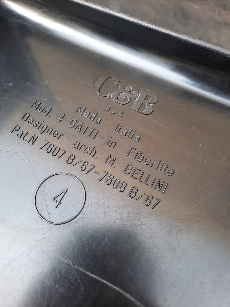 May include: Close-up of a black plastic label with white text. The label reads "C&B SpA Mod. 4 GATTI-in Fiberlite Meda Italia Designer arch. M. BELLINI Pat.N 7607 B/67-7608 B/67" and has a circle with the number "4" inside.