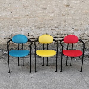 May include: Three chairs with black metal frames and colorful fabric seats. The chairs are arranged in a row on a concrete surface. The chairs are blue, yellow, and red.
