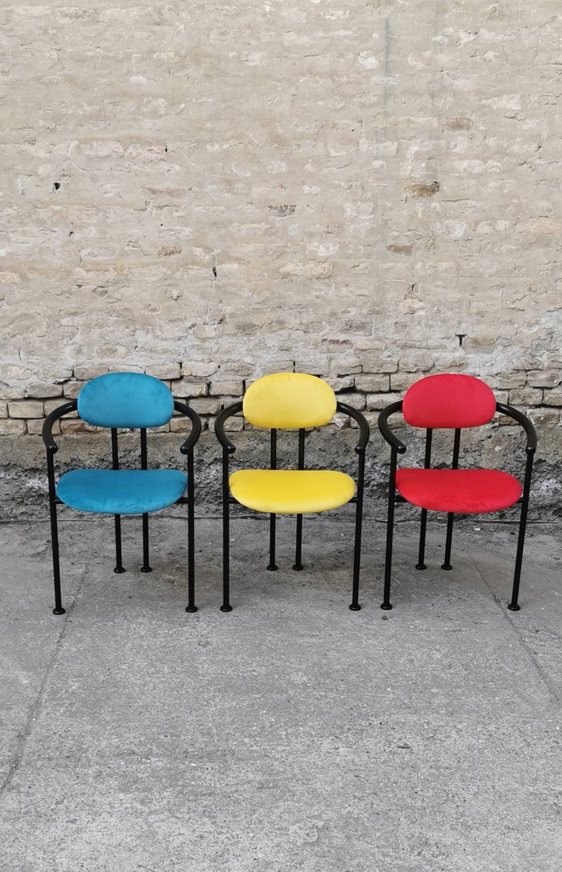 May include: Three chairs with black metal frames and colorful fabric seats. The chairs are arranged in a row on a concrete surface. The chairs are blue, yellow, and red.