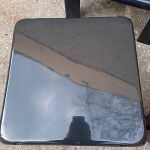 May include: A black, glossy, square-shaped side table with rounded edges. The table is made of a smooth, hard material and reflects the surrounding environment.