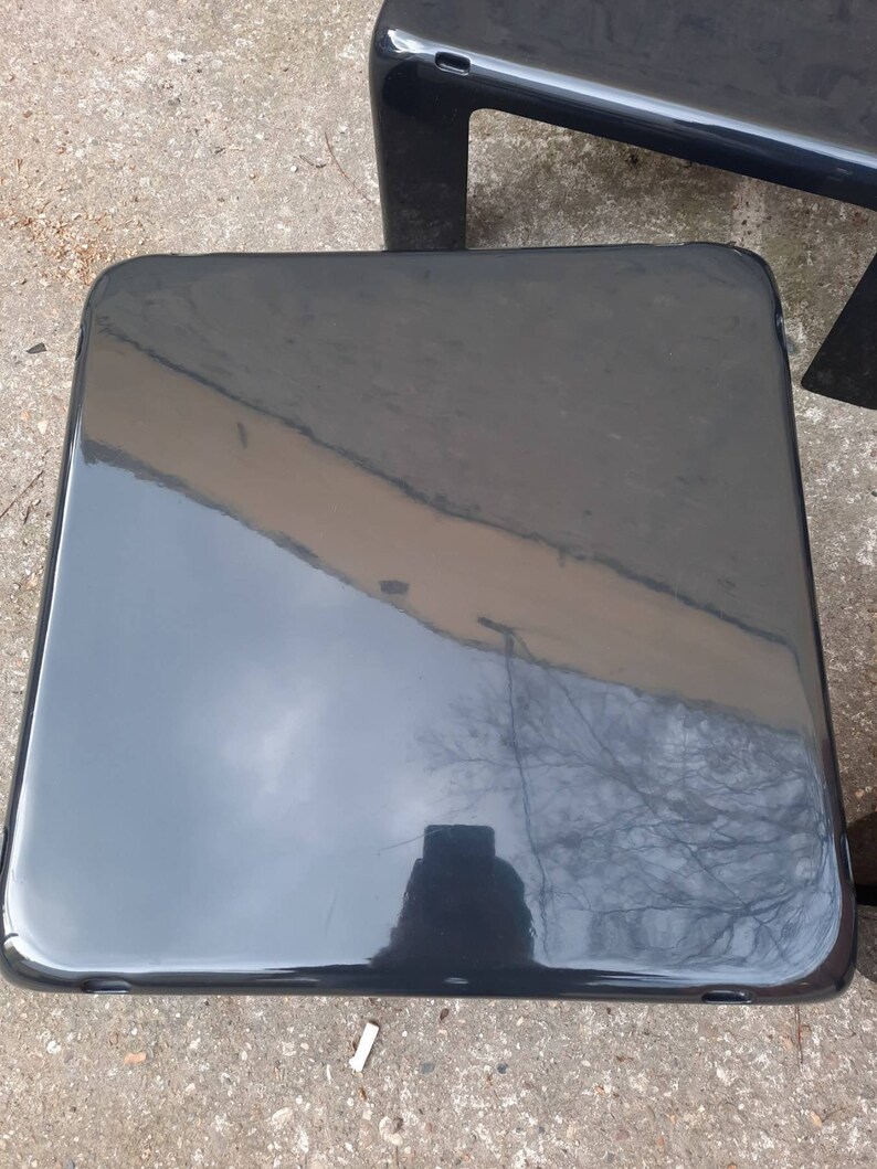 May include: A black, glossy, square-shaped side table with rounded edges. The table is made of a smooth, hard material and reflects the surrounding environment.