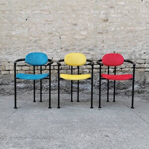 May include: Three chairs with black metal frames and colorful fabric seats. The chairs have a rounded back and are arranged in a row. The chair on the left has a blue seat, the middle chair has a yellow seat, and the chair on the right has a red seat.