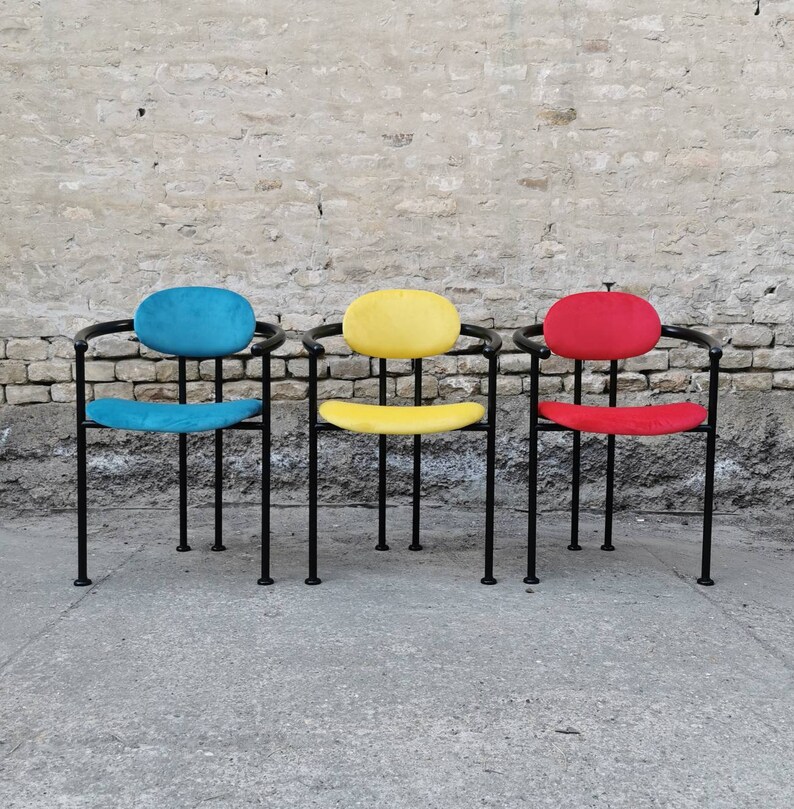 May include: Three chairs with black metal frames and colorful fabric seats. The chairs have a rounded back and are arranged in a row. The chair on the left has a blue seat, the middle chair has a yellow seat, and the chair on the right has a red seat.
