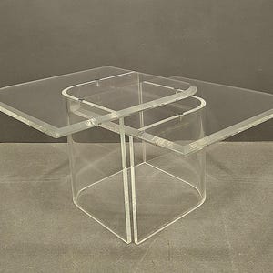 May include: Clear acrylic coffee table with a unique design. The table features a square base and a top with two overlapping rectangular sections. The edges are rounded, and the table is transparent, allowing light to pass through.