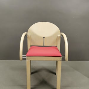 May include: A light wood chair with a red upholstered seat and curved armrests. The chair has a unique design with a cutout in the back.