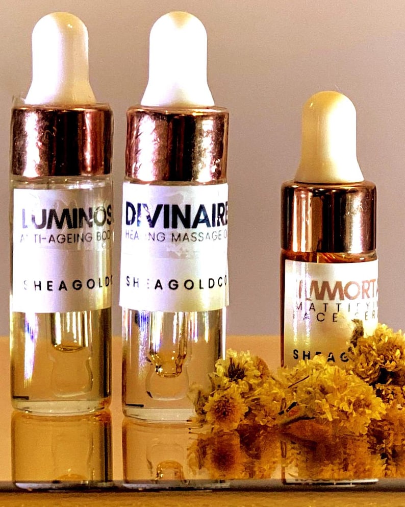 Op de afbeelding: Drie doorzichtige glazen druppelflesjes met witte doppen en metalen kragen. De etiketten vermelden LUMINOS ANTI-AGEING BODY, DIVINAIRE HEALING MASSAGE OIL en IMMORTAL MATTIFYING FACE SERUM van SHEAGOLDCO. Kleine gele gedroogde bloemen liggen aan de voorzijde.