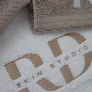 Beige embroidered towel with a large bold monogram design and small text underneath, styled on a light background for a spa or beauty business gift.