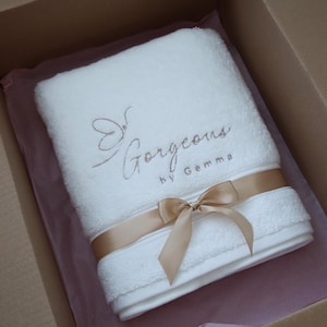 White towel neatly tied with a gold ribbon bow and presented in a gift box, showing the finished packaging of a personalised towel order.