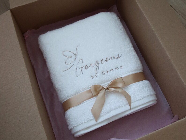 White towel neatly tied with a gold ribbon bow and presented in a gift box, showing the finished packaging of a personalised towel order.