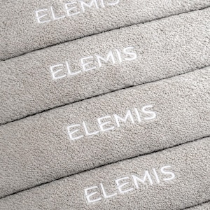 Stack of grey embroidered towels featuring small repeated lettering stitched along the folded edge, shown close-up to highlight the placement and thread detail.