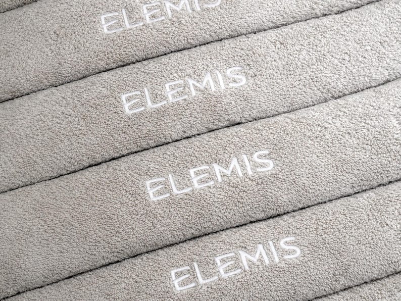 Stack of grey embroidered towels featuring small repeated lettering stitched along the folded edge, shown close-up to highlight the placement and thread detail.