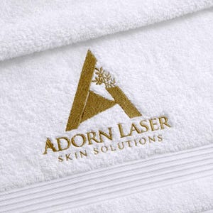 White towel featuring an elegant triangular monogram emblem embroidered in gold thread, a refined laser-cut style logo design.